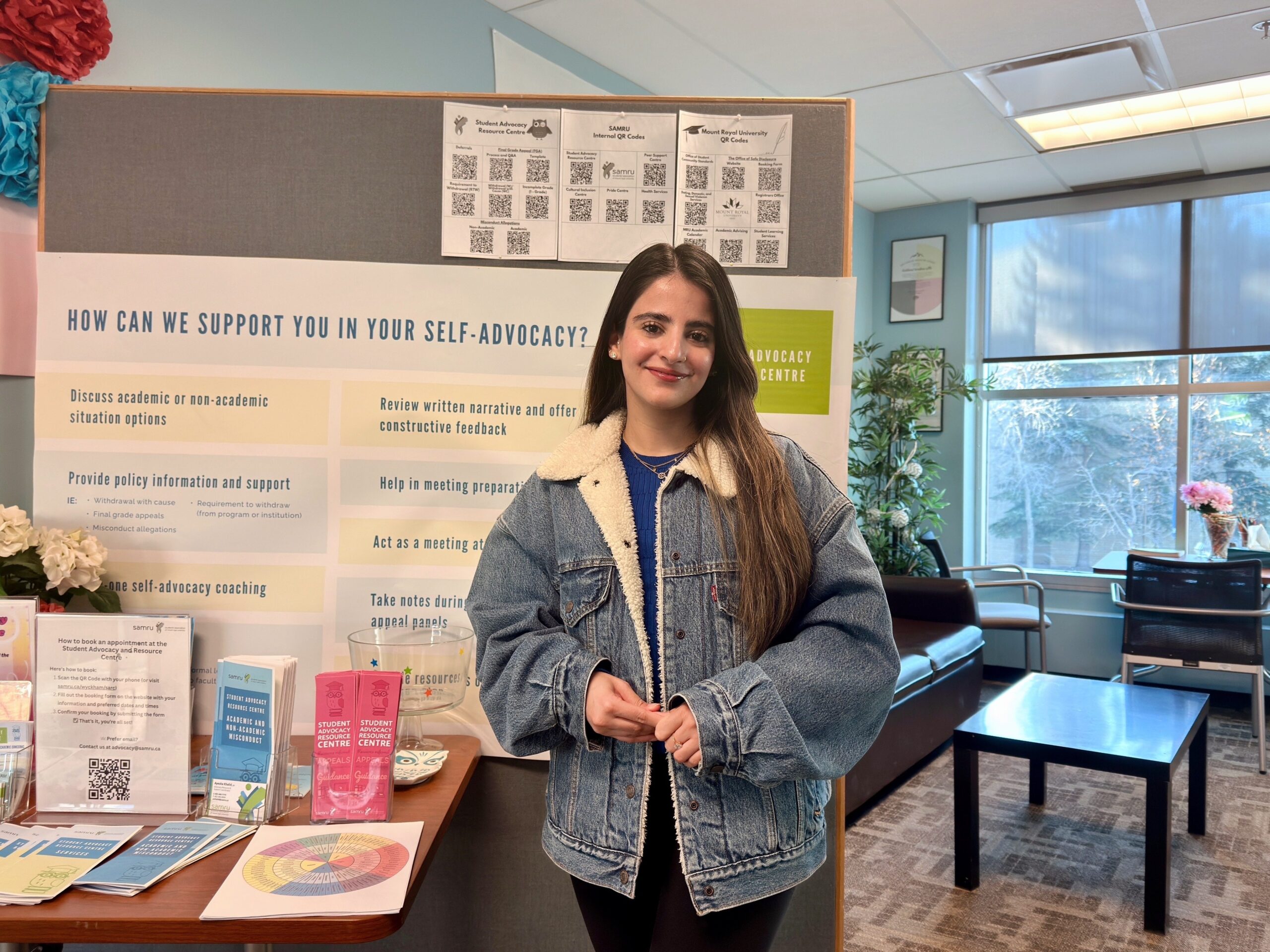 SARC Coordinator Ayesha standing in the Student Advocacy Resource Centre in front of a board of resources. 